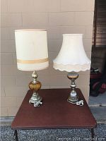 Both retro lamps displayed side by side on a wooden table outdoors against a beige wall, showing different lampshade shapes and base materials.
