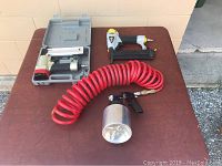 Photo of air tools on a brown table outside against beige wall including pneumatic nailers, red coiled air hose, and spray gun with container.
