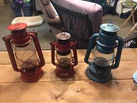 Three vintage oil lamps of different sizes and colors on a wooden table in an indoor setting.