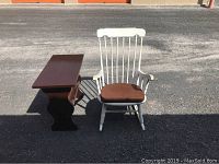 Photo showing the white wooden rocking chair with brown cushion beside the dark wooden side table on outdoor pavement.