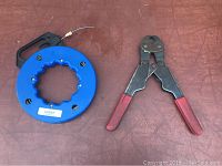 Photo showing the blue fish tape reel and Pex crimper tool with red handles on a brown surface.