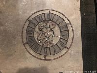 Large round wall clock with Roman numerals and mesh background showing visible gears in the center, worn rustic finish.