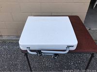 Closed white Thule camping table with handle lying on a brown table, showing Thule branding.