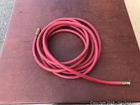 Coiled red air hose displayed on a dark reddish-brown surface, showing metal connectors on both ends.