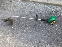 Full view of green FeatherLite gas weed eater with curved shaft and protective guard laying on the ground.