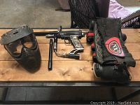 Paintball mask, marker, two hoppers, spare barrel, and harness arranged on a wooden picnic table.