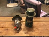 Photo of two camping lanterns on a wooden table: one green pump-style and one propane lantern with glass globe and red knob