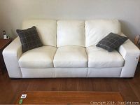 Front-facing view of the white leather sofa with two gray grid-pattern throw pillows on hardwood floor