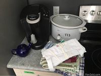 View of Keurig coffee maker, blue ceramic teapot, Betty Crocker crock pot, and folded assorted tea towels on kitchen counter.