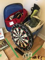 Overall lot including dart board leaning on two Rubbermaid backpacks filled with various items including a folded travel blanket and a blow up mattress.