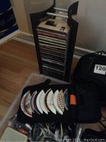 Photo showing a black CD holder with multiple discs inserted and loose CDs beside it, assorted cords in plastic container below.