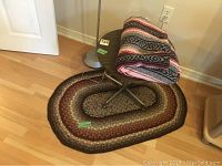 Photo shows floor lamp base partially visible behind a round wooden table with a folded multicolored woven blanket on top, and an oval braided floor mat underneath.