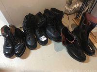Four pairs of men's boots arranged side by side on floor including black steel toe boots, rubber galoshes and formal boots.