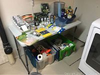 Full view of folding utility table with assorted household items and supplies on and below it, including plastic containers, flashlight, pylons, and other tools.