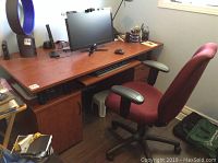 View of the wooden desk with a computer monitor, keyboard tray pulled out, office chair in front, and task lamp on desk.