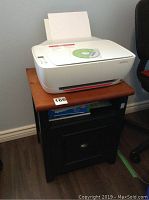 White HP DeskJet 3634 printer placed on wooden printer stand with black body and natural wood top, numbered sticker on stand.