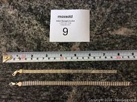 Two silver bracelets laid flat on a dark grey surface next to a measuring tape showing lengths around 18 and 19 cm.