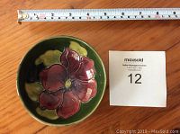 Top view of round dish showing large red flower and green background with measuring tape showing approx. 5 inches diameter.