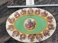 Top view showing entire oval porcelain platter with painted romantic couples in multiple vignettes around the rim and center, green and gold background, 24kt gold trim on edge, ruler for scale showing about 13 inches length.