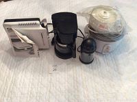 Lot of small kitchen appliances including coffee maker, sandwich grill, citrus juicer, rice cooker, and cheese grater arranged on a white surface.