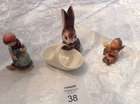 Three porcelain figurines arranged on a lace tablecloth, including a rabbit basket and two child figures.
