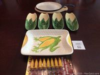 Four small ceramic corn husk-shaped dishes, rectangular serving dish with raised corn decoration, and yellow corn holders on skewers.