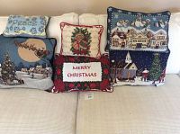 Six decorative Christmas pillows arranged on a white couch, showing a variety of festive designs including snowy village scenes, Santa and reindeer, text motifs, and holiday symbols.