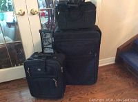 Photo of three black luggage pieces: large Atlantic suitcase, Tracker carry-on, and tote bag, standing on wood floor.