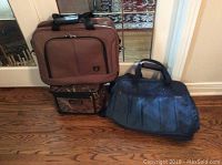 Three luggage items including a brown wheeled carryon bag, a blue nylon bag, and a small toiletry case placed on a wooden floor.
