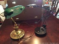 Two desk lamps on table, showing vintage green banker's style lamp with brass base and modern black adjustable lamp with clamp base.