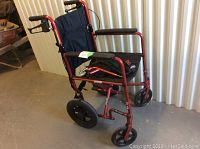Red lightweight transport wheelchair with black seat, armrests, and rear push handles with brakes, photographed from side angle to show overall condition.