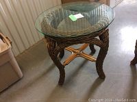 Round wicker end table with clear glass top, legs curved and woven with a cross brace beneath.