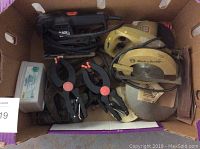 Top-down image of a cardboard box containing a Black & Decker circular saw, a small sander, several black and red spring clamps, and another electric tool with a power cord.