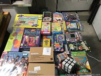 Overview photo of the assorted new games in original packaging arranged on a table including board games, card games, handheld electronic games, and puzzles.