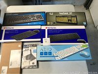 Photo showing seven boxed and loose keyboards from various brands including Verbatim, AZIO, Adesso, and Kensington, as well as a document holder, arranged on a flat surface.