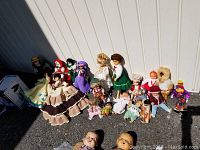 Wide view showing multiple dolls of various types and sizes arranged together outdoors on asphalt against a wall, including crocheted and plastic dolls in various dresses.