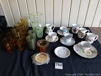 Lot display of assorted china cups, saucers and coloured glasses on a black cloth with wall background.