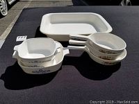 Seven assorted baking dishes displayed on a black surface including two white CorningWare dishes with blue floral patterns, three white CorningWare skillets with brown floral patterns on the handles, and one rectangular enamelled metal baking dish.