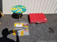 Image showing vintage round metal table with floral top and original box, alongside a red vintage Samsonite suitcase on pavement.