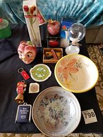 Overview of various decorative and collectible items displayed on a black cloth including ceramic apple figurines, glass hourglass, ceramic bowl and plate, keychains, and other small items.