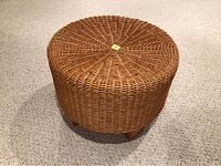 Front side angle view of the round wicker ottoman with four wooden legs on carpeted floor.