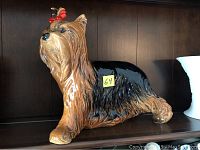 Full side view of the Goebel ceramic dog figurine on a shelf, showing the brown and black painted fur with a red bow on its head and a yellow lot number label on the side.
