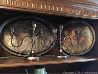 Two silver-plated candelabras displayed side by side with a large oval silver tray behind them on a wooden shelf.