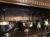 Six silver plate serving pieces including two large bowls, two small creamers, and two trays on a wood shelf