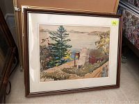 Framed watercolour painting showing a lakeside scene with rustic cabin, stone chimney, trees, and hills in the background.