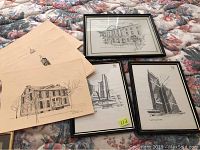 Photo showing three framed black and white cityscape sketches and four loose sketches on beige paper, arranged on a floral fabric surface.