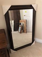 Full view of the large mirror leaning against a wall showing the reflective surface and dark wood frame with protective cardboard corners.
