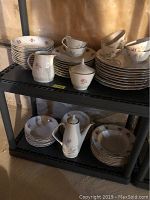 Photo showing stacked plates, bowls, cups, creamer, sugar bowl all with pink floral design and gold rim on black shelving rack.