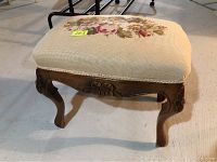 Front view of needlepoint footstool showing floral upholstery on beige fabric and carved wooden legs with floral details.