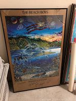 Framed signed Beach Boys Summer In Paradise poster leaning against wall, showing full image and damaged glass.